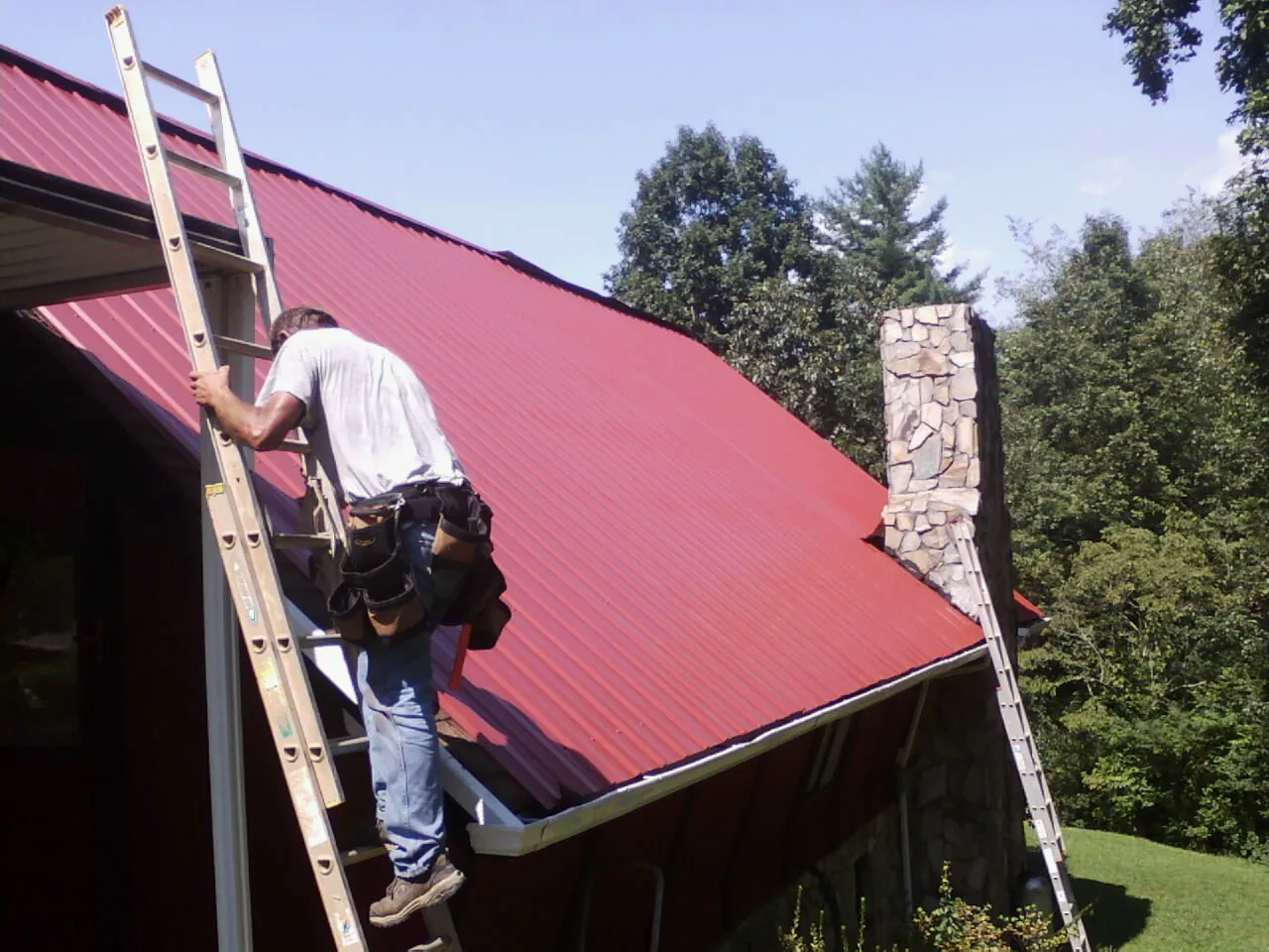 Roofer on a ladder beside a red metal roof and stone chimney — Soffit & Fascia Repair by Roofing Brothers in Phenix City